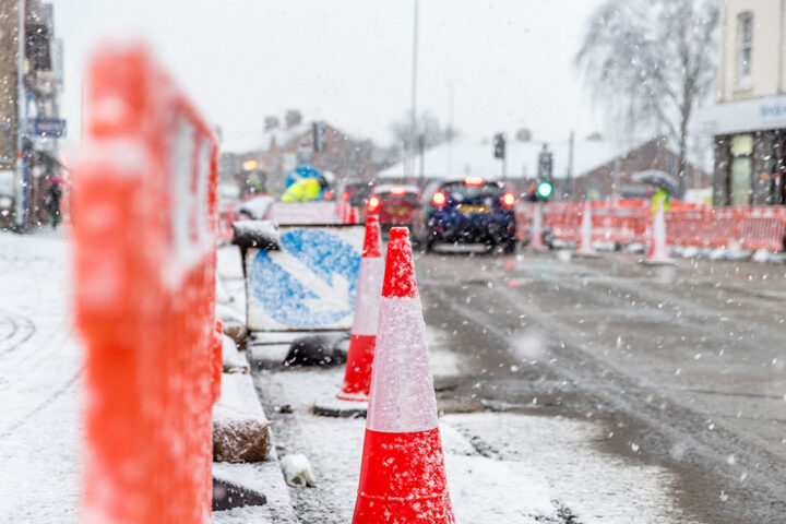 Traffic cones and construction barriers on a snowy street, with cars in the distance driving between roadworks