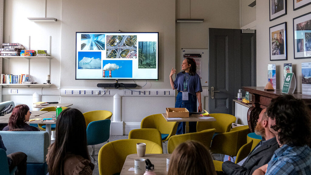 A person wearing a blue tshirt and denim skirt tsand next to a presentation screen, gesturing with their hand. They are in a room with several other people, who are sat in blue and green chairs, watching the person give the presentation.