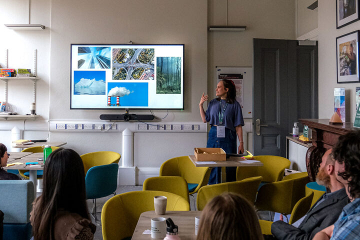 A person wearing a blue tshirt and denim skirt tsand next to a presentation screen, gesturing with their hand. They are in a room with several other people, who are sat in blue and green chairs, watching the person give the presentation.