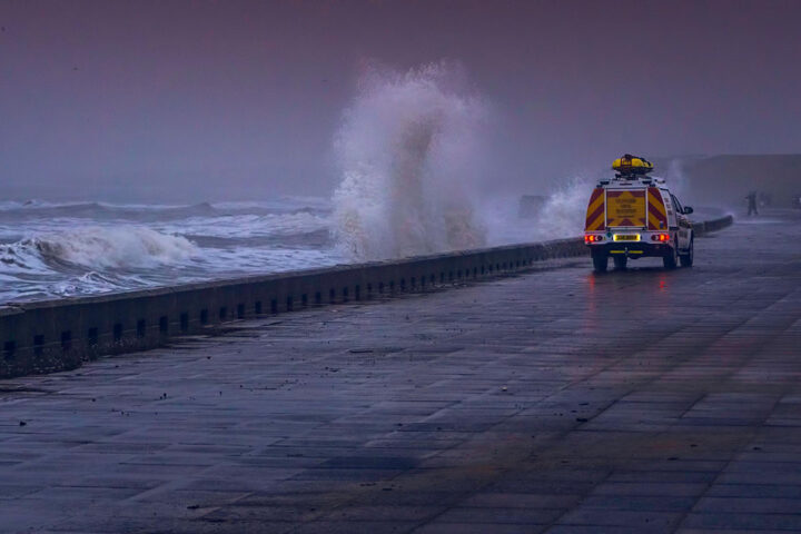 Sea water hitting spraying on coastal wall, with an orange emergency vehicle driving along