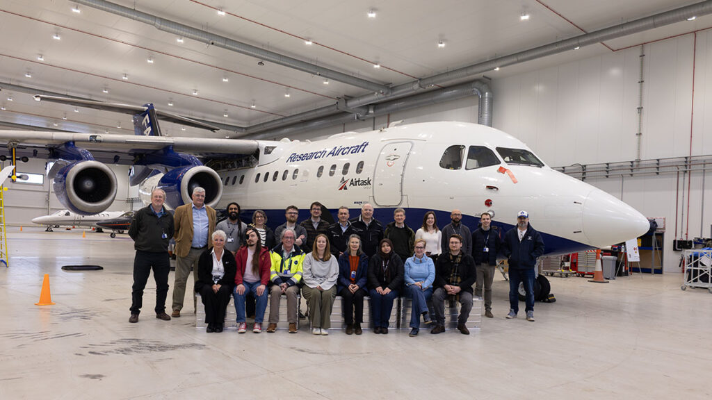 A group of people sit and stand in front of an aircraft in a hangar.