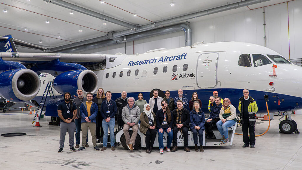 A group of people sit and stand in front of an aircraft in a hangar.
