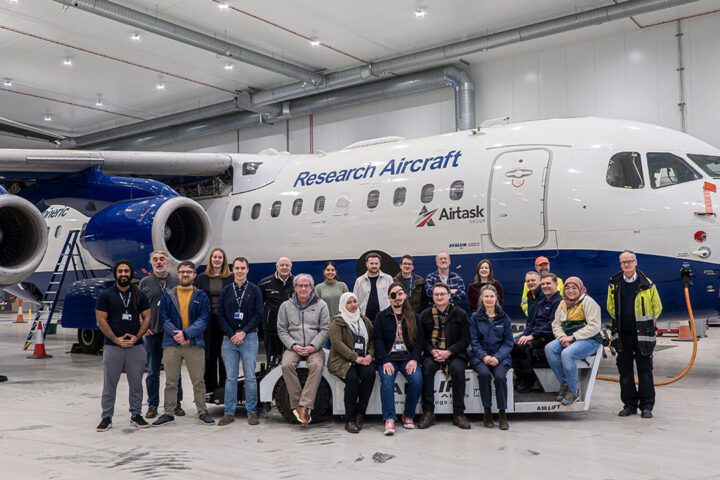 A group of people sit and stand in front of an aircraft in a hangar.