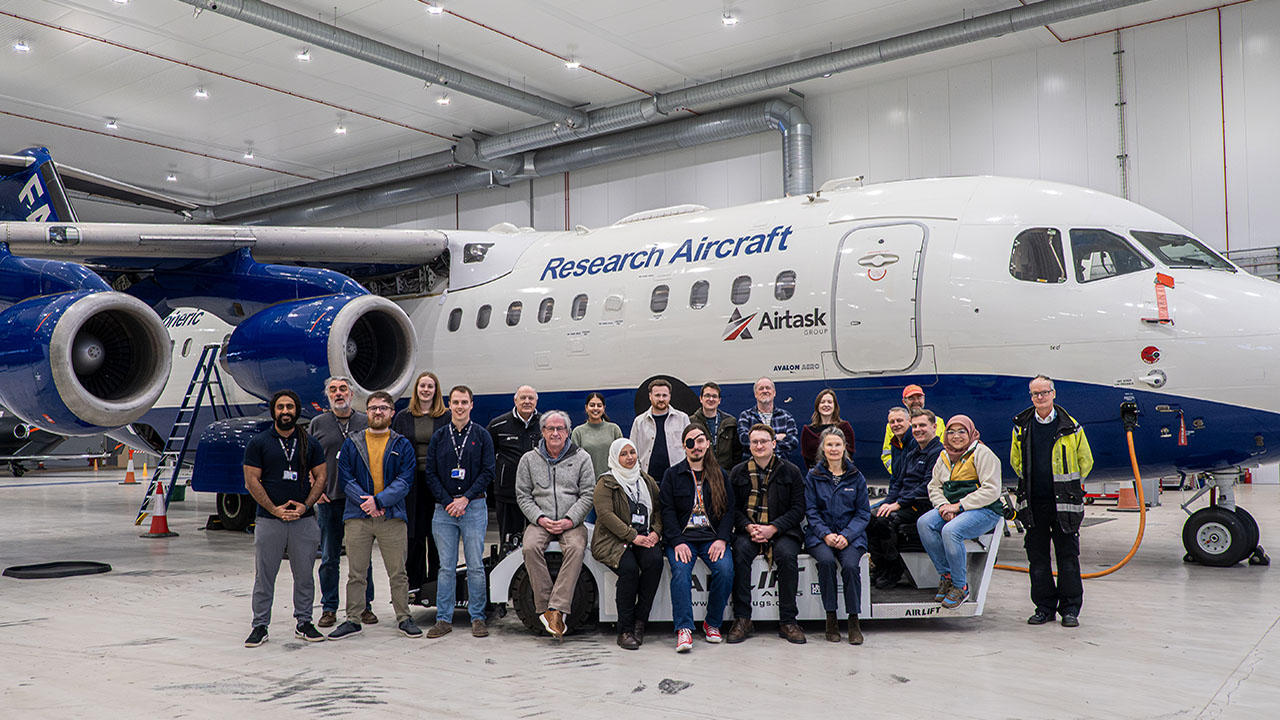 A group of people sit and stand in front of an aircraft in a hangar.