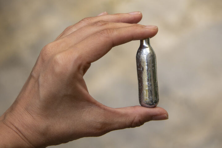 A hand holding a silver canister between their thumb and first finger.