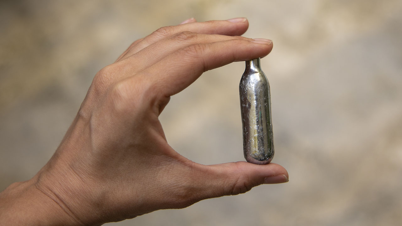 A hand holding a silver canister between their thumb and first finger.