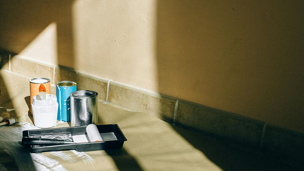 Tins of paint and decorating equipment on the floor, in front of a beige wall. The light from a nearby window is cast onto the wall.