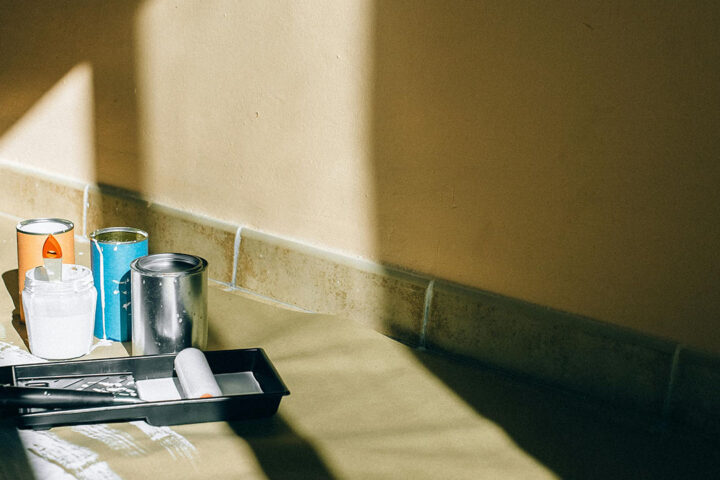 Tins of paint and decorating equipment on the floor, in front of a beige wall. The light from a nearby window is cast onto the wall.