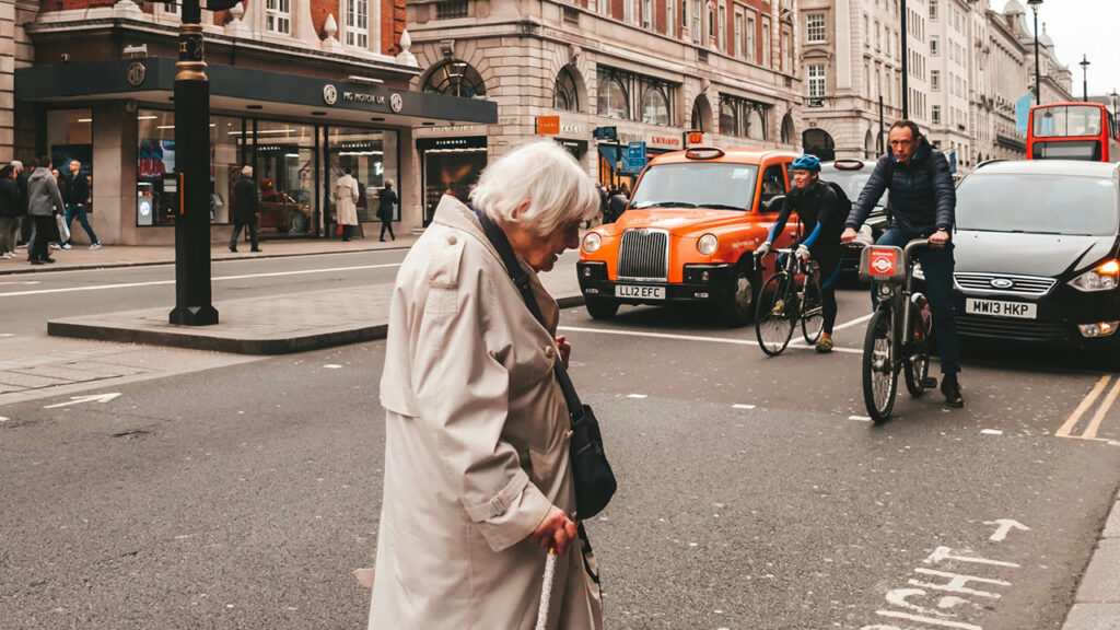 Older person wearing a beige coat and using a walking stick crosses the road in front of queued up traffic.