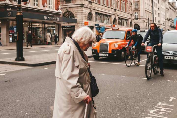 Older person wearing a beige coat and using a walking stick crosses the road in front of queued up traffic.