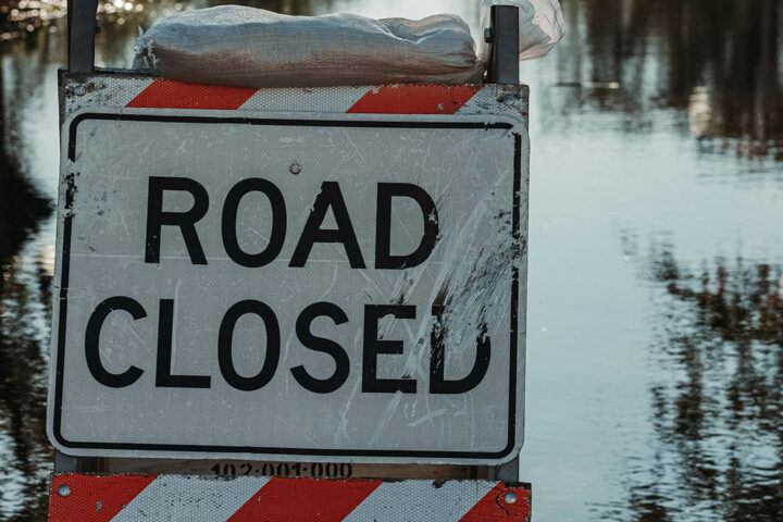 Black, white and red sign that says road closed, in front of still flood water on a tree lined road.
