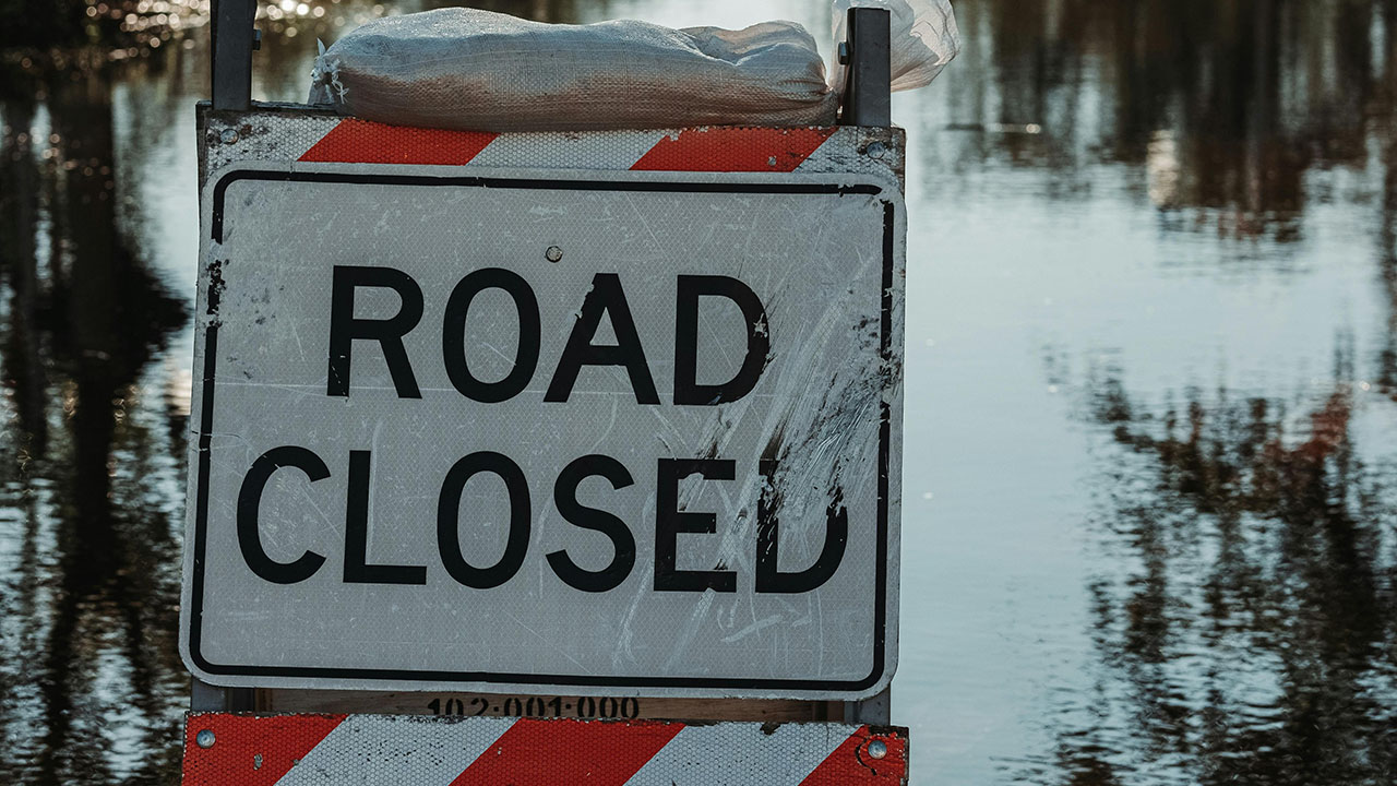 Black, white and red sign that says road closed, in front of still flood water on a tree lined road.