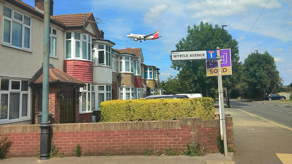 An aircraft flies low over trees and houses in a residential area.