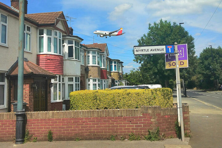 An aircraft flies low over trees and houses in a residential area.