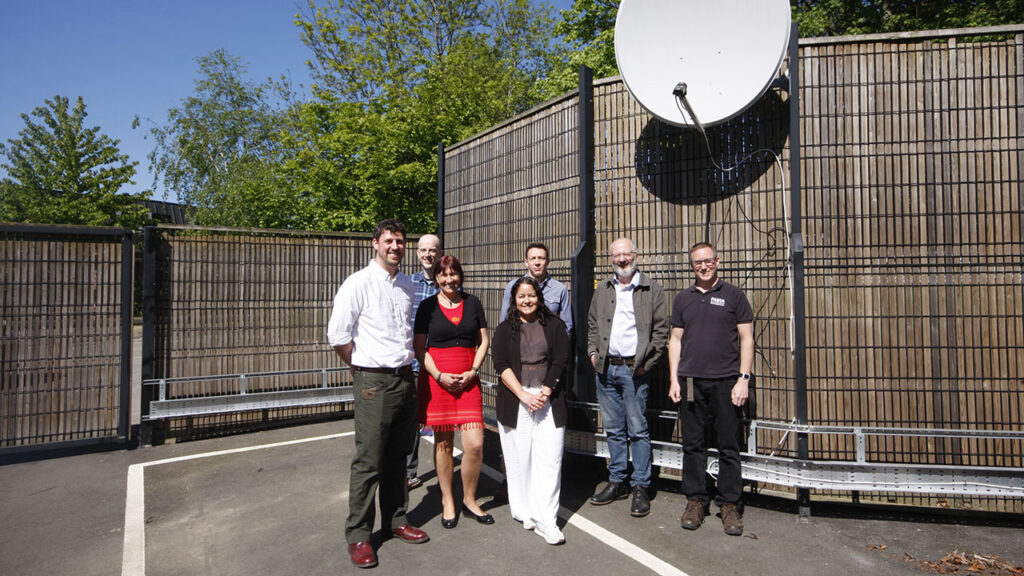 Group of 6 smiley people stood underneath a pale grey satellite dish, in a fenced compound.