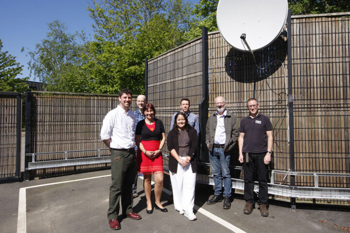 Group of 6 smiley people stood underneath a pale grey satellite dish, in a fenced compound.