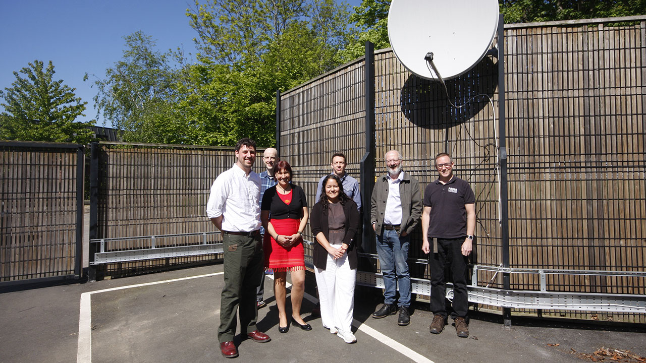 Group of 6 smiley people stood underneath a pale grey satellite dish, in a fenced compound.