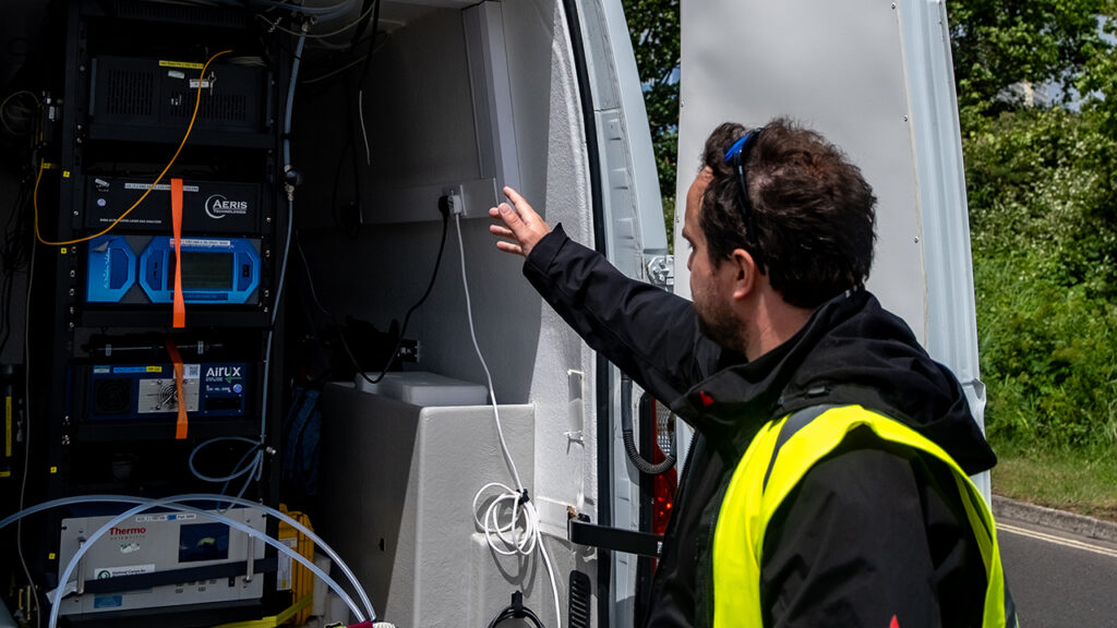 A person wearing a high visibility jacket points their arm towards the back of an open van. In the van there are stacks of air quality monitoring equipment.