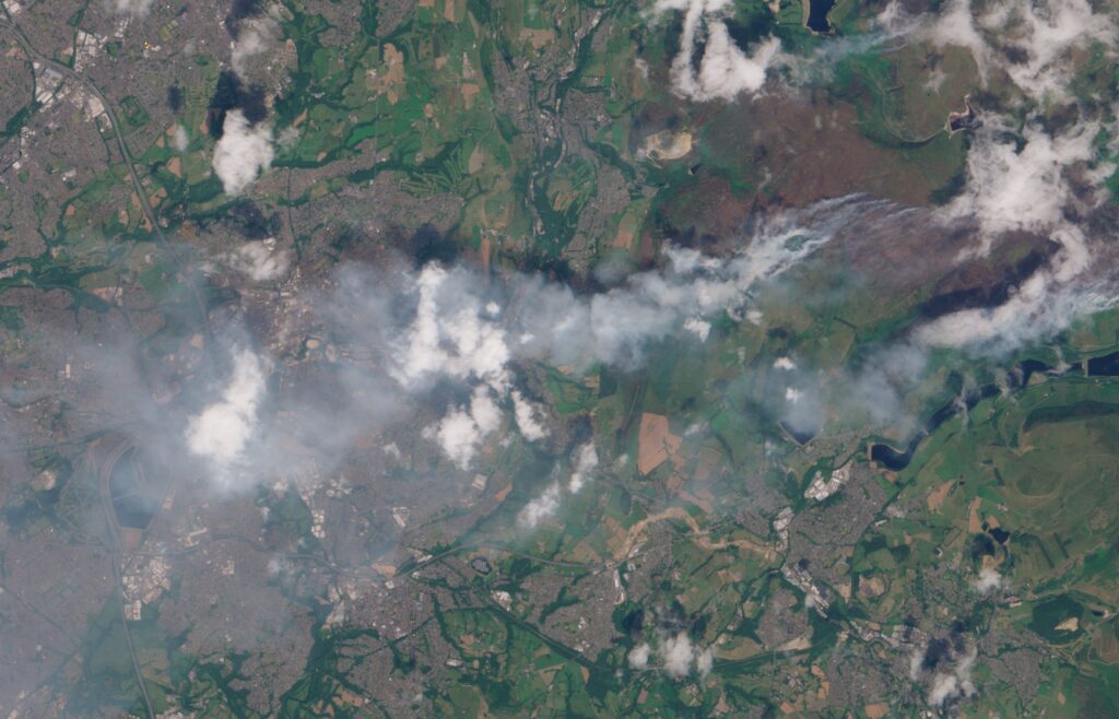A satellite image showing wildfire smoke blowing towards the west across green and brown land towards a more urban area.