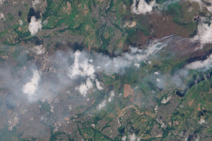 A satellite image showing wildfire smoke blowing towards the west across green and brown land towards a more urban area.
