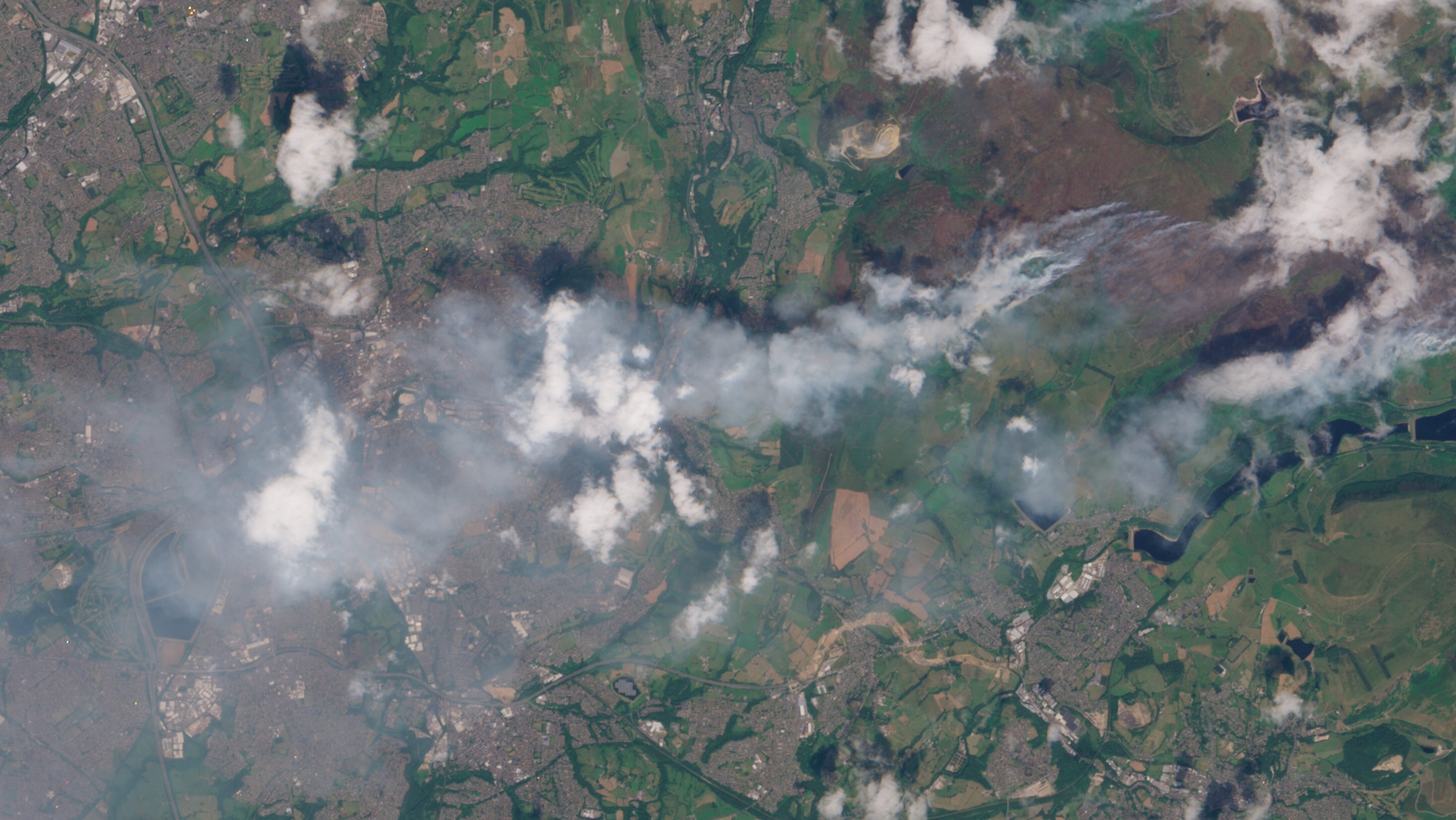 A satellite image showing wildfire smoke blowing towards the west across green and brown land towards a more urban area.