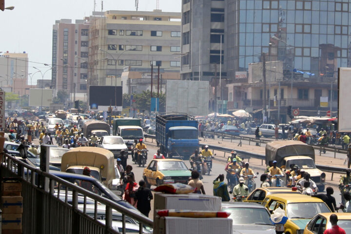 Busy street with lots of people walking, riding scooters, and driving cars and trucks.