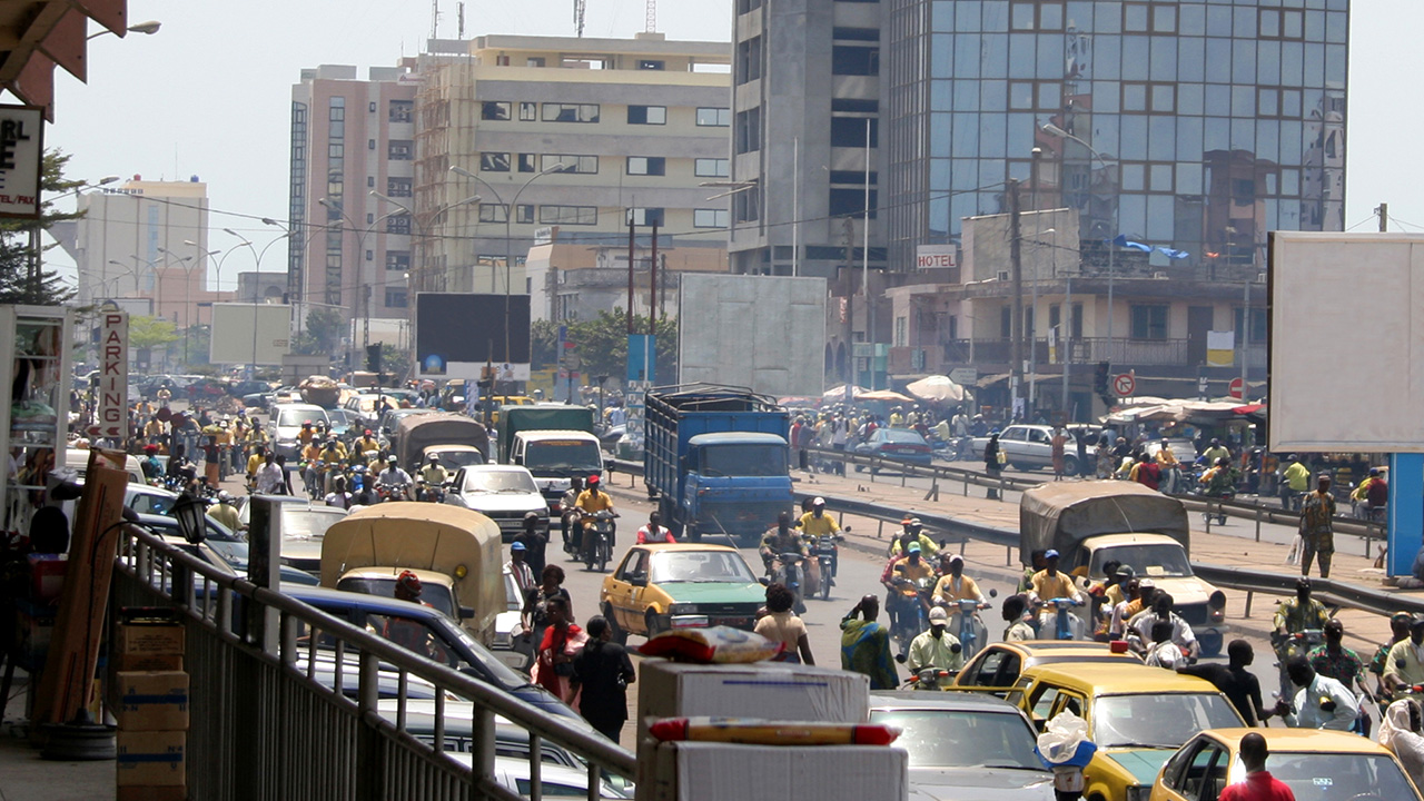 Busy street with lots of people walking, riding scooters, and driving cars and trucks.