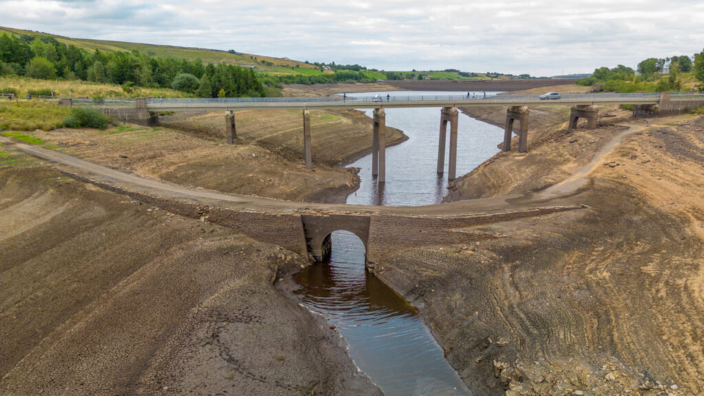 Low water levels in a very dry and brown reservoir, with 2 bridge crossings, and some people on one bridge looking down at the exposed ground.