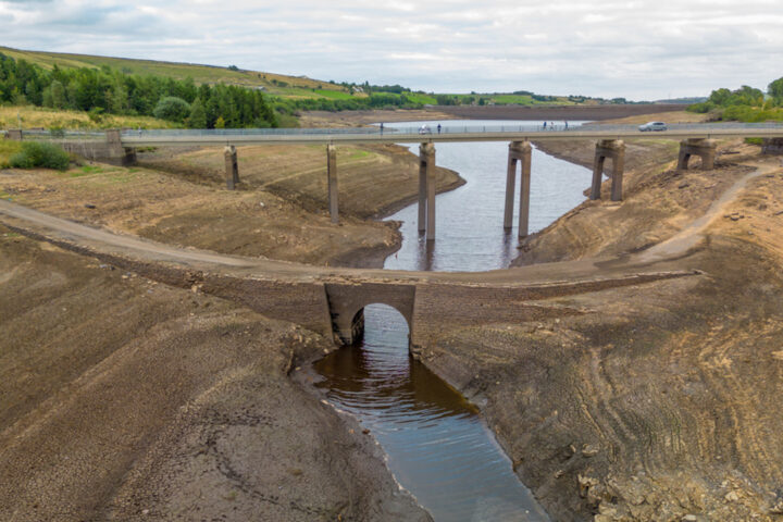 Low water levels in a very dry and brown reservoir, with 2 bridge crossings, and some people on one bridge looking down at the exposed ground.