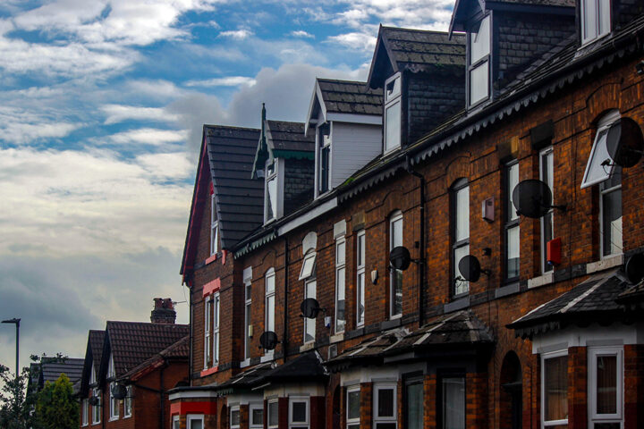 A row of terraced houses in Greater Manchester. There is a blue sky with white clouds in the background. Some houses have their windows open.