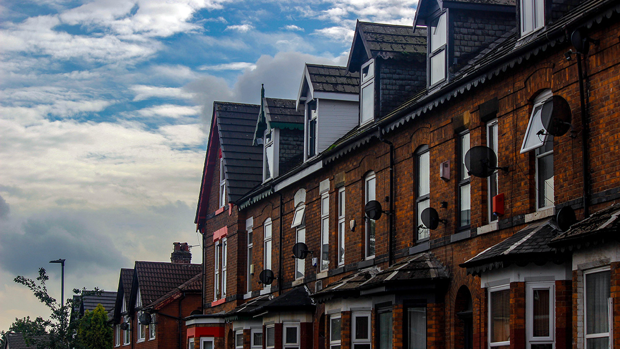 A row of terraced houses in Greater Manchester. There is a blue sky with white clouds in the background. Some houses have their windows open.