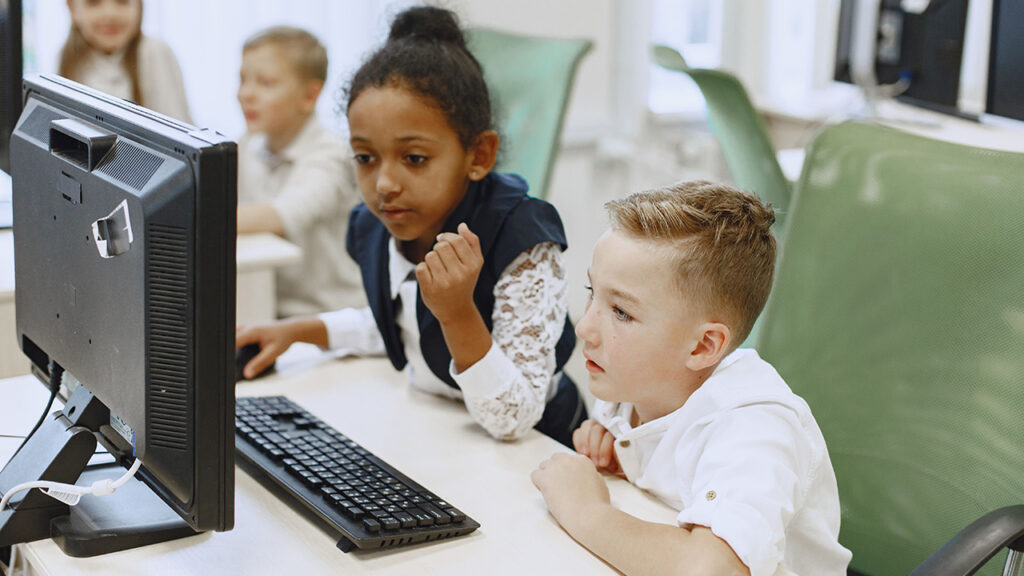 Two children sat on green chairs at a desk, sharing the use of a desktop computer.