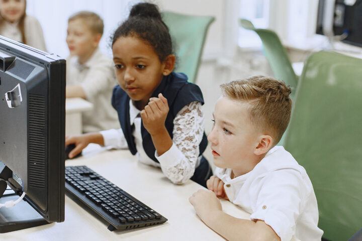 Two children sat on green chairs at a desk, sharing the use of a desktop computer.