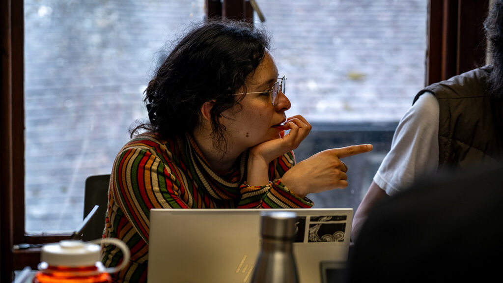 A person wearing glasses is sat in front of a laptop, and points with their index finger to  one side.