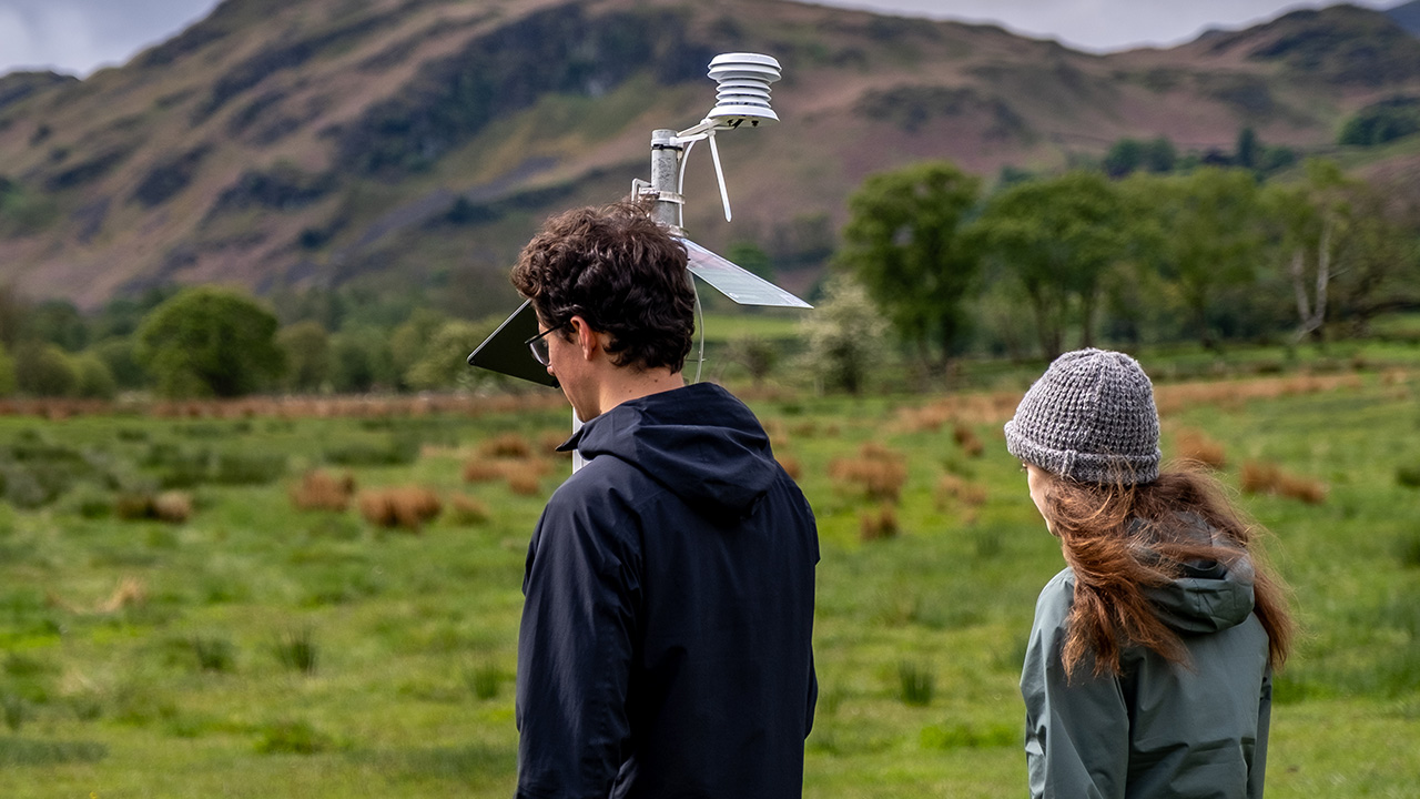 Two people walking in a field, with mountains in the distance. One person carries a small mobile weather station.