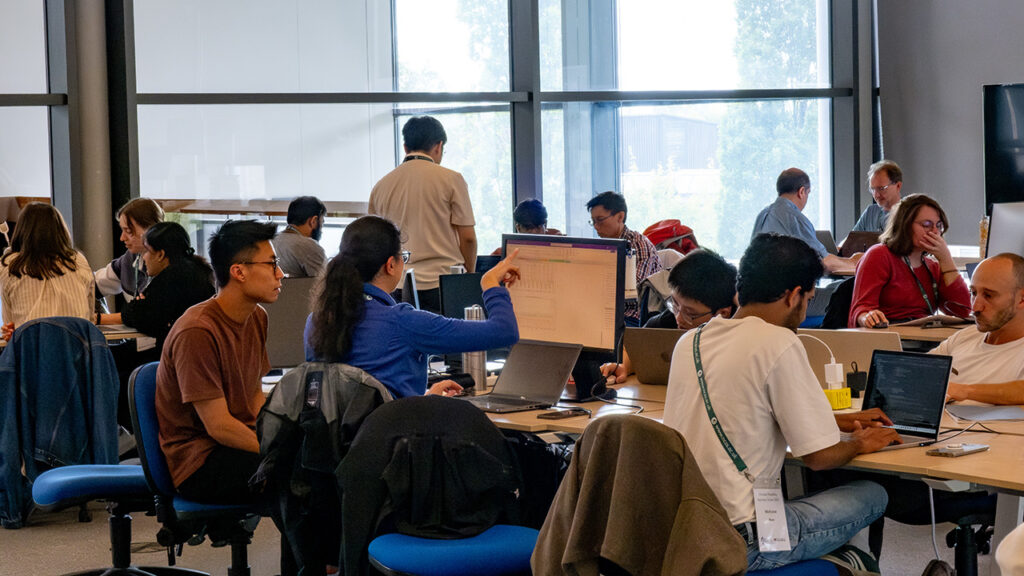 People sat on blue desk chairs at office desks, in groups or on their own, working on laptops.