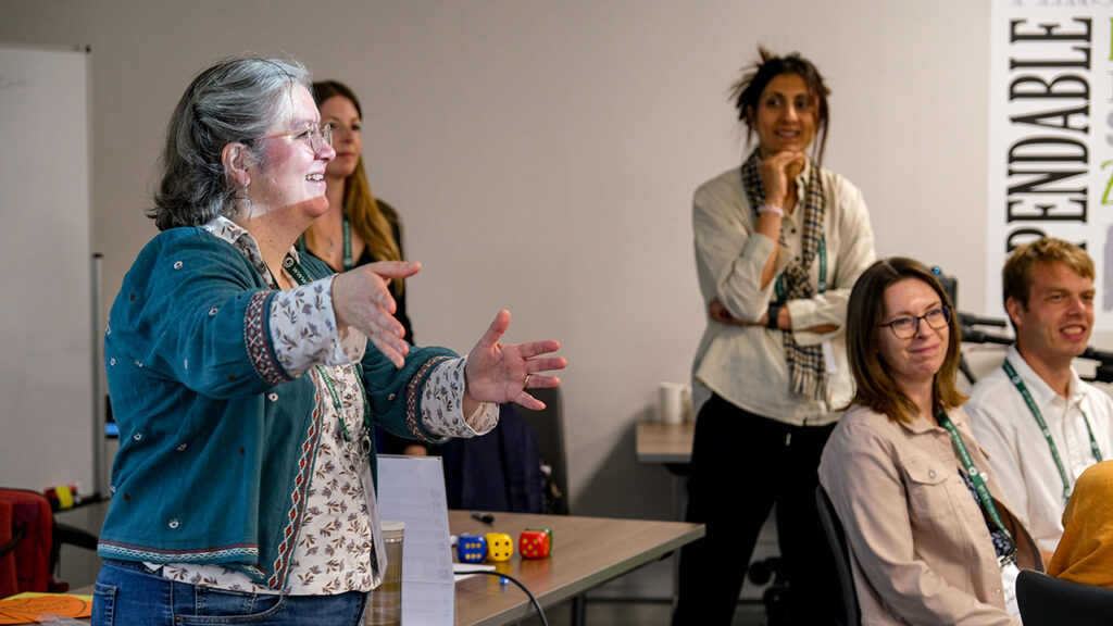 A person standing up and lifting their arms while speaking. They are in a room with other people, some are stood and some are sat on chairs.