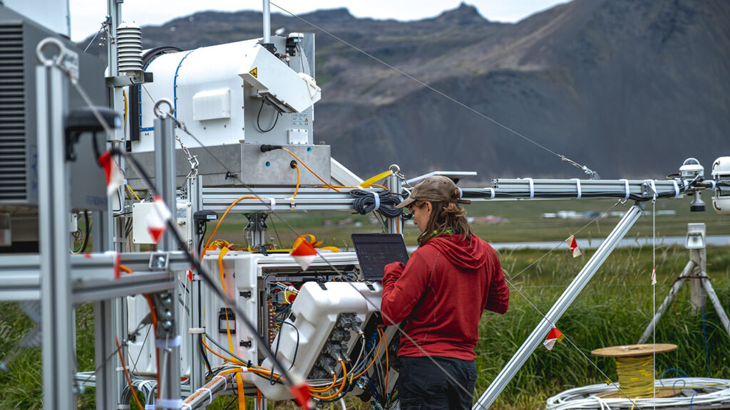 A person wearing a cap and a red jumper is working at a laptop, which is balanced on a scientific instrument. There are metal structures and cables in view. In the background there is green land and mountains.