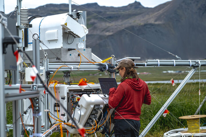 A person wearing a cap and a red jumper is working at a laptop, which is balanced on a scientific instrument. There are metal structures and cables in view. In the background there is green land and mountains.