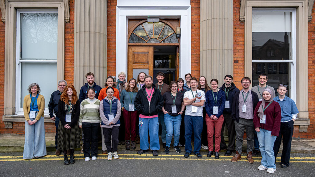 Group of people stood together in front of the door to a big red brick building with stone columns.