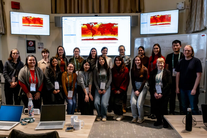 A group of people stand underneath 3 large screens displaying atmospheric data in a red global view graphic.