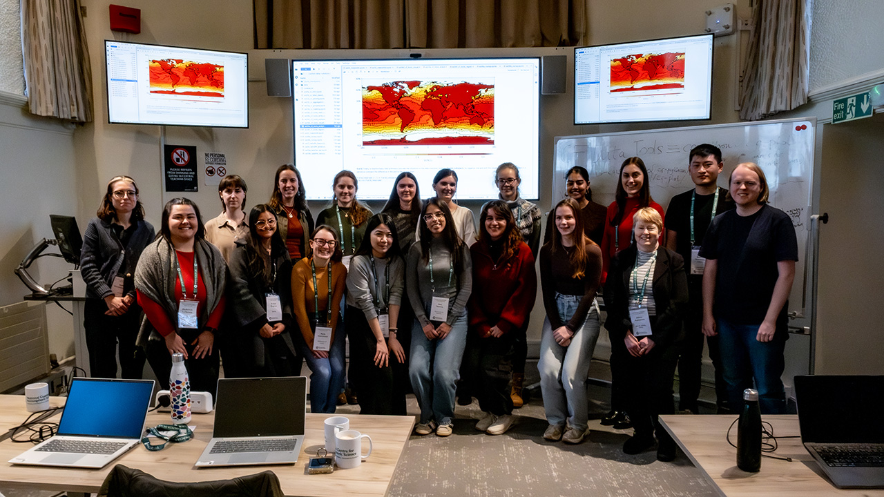 A group of people stand underneath 3 large screens displaying atmospheric data in a red global view graphic.