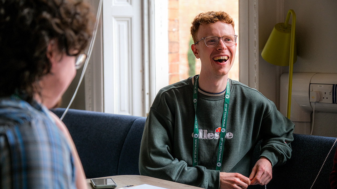 A person laughing, sat across from someone else at a table.