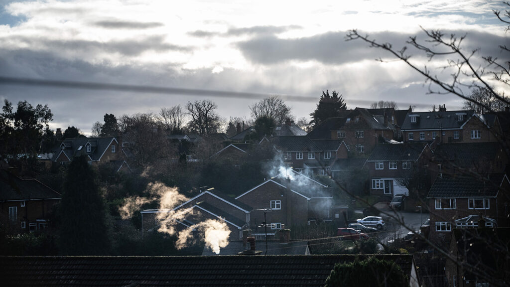 Houses and parked cars in a residential area. There is sun shining through grey clouds in the sky. There is smoke rising from chimneys.