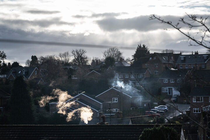 Houses and parked cars in a residential area. There is sun shining through grey clouds in the sky. There is smoke rising from chimneys.