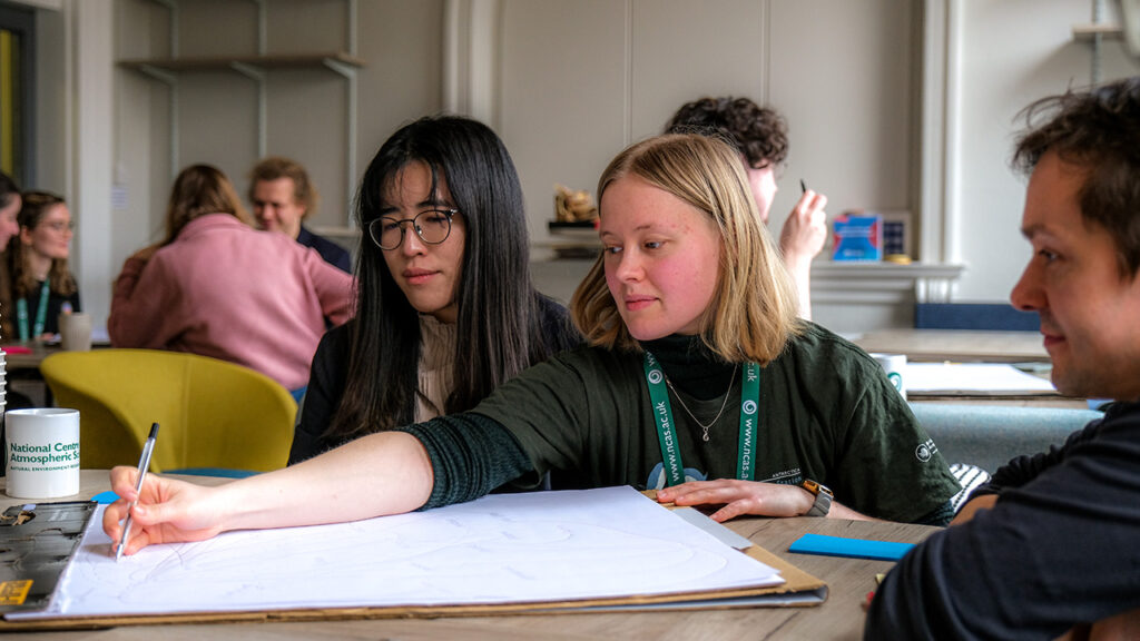 One person in a small group of people sat at a table draws on a large piece of paper. Thera re other people doing the same in the background, on other tables.