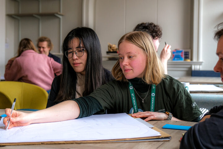 One person in a small group of people sat at a table draws on a large piece of paper. Thera re other people doing the same in the background, on other tables.