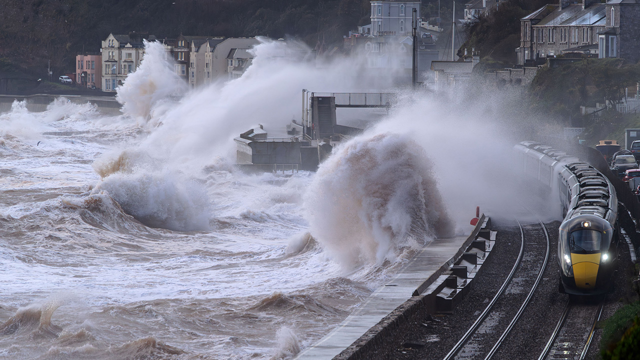 Waves from the sea splashing over a moving train, in front of seaside houses.